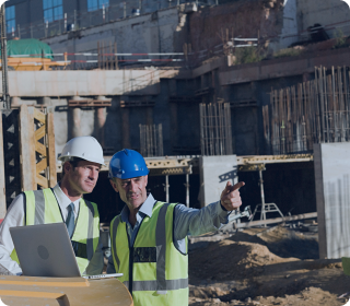 Engineers wearing safety helmets and vests supervising construction site with laptop and building framework in the background