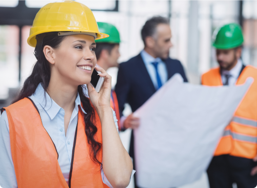 Female construction engineer wearing a safety helmet and vest, talking on the phone at a building site with colleagues reviewing blueprints in the background